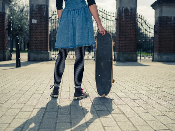 Young woman standing at gates of park with skateboard — Stock Photo, Image