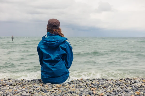 Kvinnan i blå jacka sitter på stranden — Stockfoto