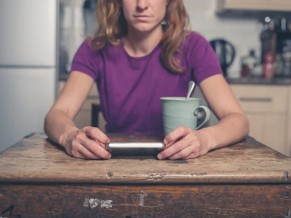 Jeune femme dans la cuisine avec thé et téléphone intelligent — Photo