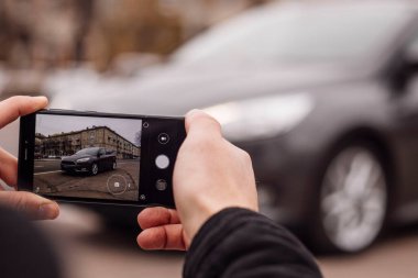 Bir adam şehrin ortasında telefonla bir arabanın fotoğrafını çekiyor.