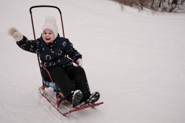 Bir çocuk kışın karda kızağa biner ve elini sallar.