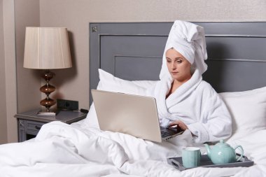 Yatakta dizüstü bilgisayarı olan bir kadın. Banyo yaptıktan sonra otelde dinleniyor.