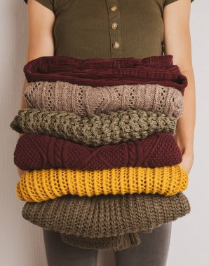 Genç bir kadın elinde sonbahar renklerinde kalın giysiler tutuyor. Sarı, koyu yeşil, kahverengi kazaklar.