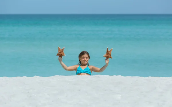 Pequena menina sorridente alegre sentado na areia branca praia tropical e segurando estrela do mar — Fotografia de Stock