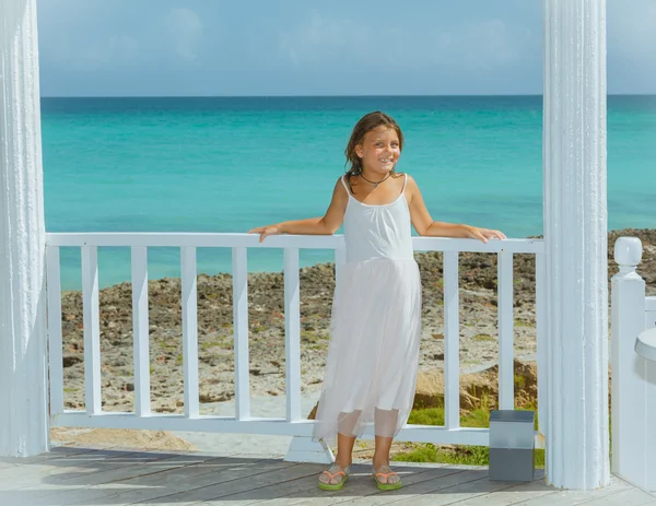 Smiling happy little girl in white light dress standing against tranquil azure ocean background — Stock Photo, Image