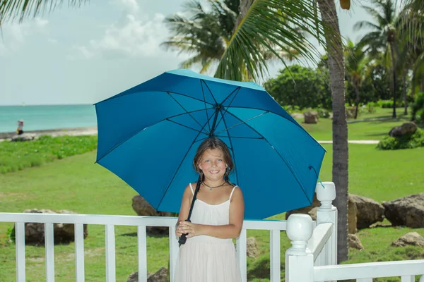 Happy little girl standing with big blue umbrella against natural tropical background — Stock Photo, Image