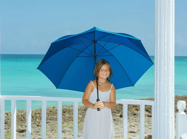 joyful little girl holding fashioned , big blue umbrella and leaning against white wooden fence on tropical rocky seashore and ocean background