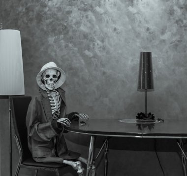 Happy skeleton sitting behind the table in dark dramatic room environment, wearing leather jacket and white hat and waiting for Halloween party clipart