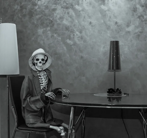 Esqueleto feliz sentado atrás da mesa em ambiente de quarto dramático escuro, vestindo jaqueta de couro e chapéu branco e esperando pela festa de Halloween — Fotografia de Stock