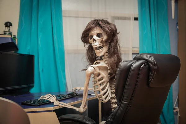 Smiling, crazy skeleton in a wig sitting in room on black leather chair behind the desktop — Stock Photo, Image