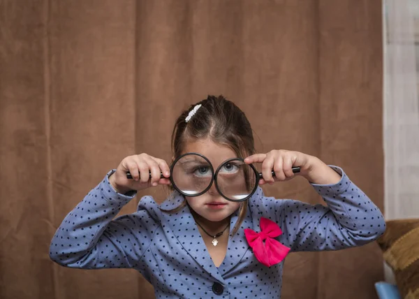 Şık şık komik küçük kız yalak doğru büyütülmüş gözlük kahverengi perdeleri arka plan üzerinde seyir. — Stok fotoğraf