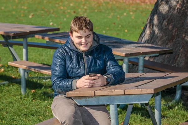 Joyful tonårspojke sitter avkopplande i höst park och skickar SMS på mobiltelefon — Stockfoto