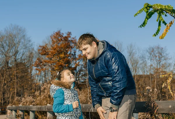 Brother sharing the headphones with little joyful sister and listening funny music in autumn park on sunny beautiful day — Stock Photo, Image