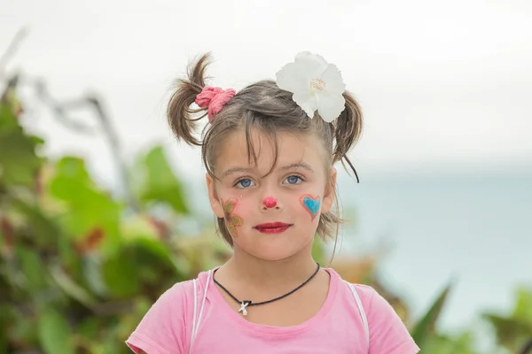 Jouful meisje met verrast reactie op haar geschilderde gezicht permanent in de buurt van het strand tegen de Oceaan en tropische tuin achtergrond — Stockfoto