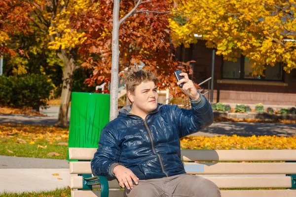 Hamdsome teenage boy  sitting on the bench in  park and making self photograph on his cellphone — Stock Photo, Image