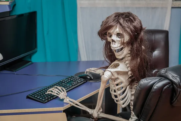 Joyful smiling skeleton in a wig sitting in chair behind the desktop computer — Stock Photo, Image