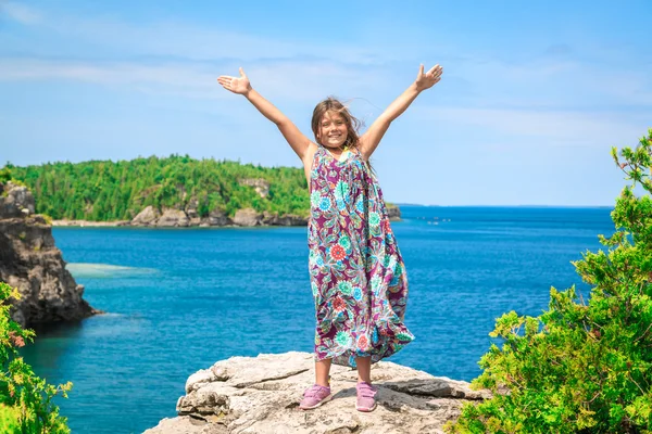 Happy, joyful child girl standing with hands up on the edge of high cliff above gorgeous amazing Cyprus lake — Stock Photo, Image
