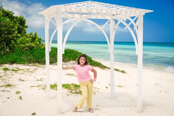 Фешенебельная улыбающаяся маленькая девочка, стоящая в беседке под открытым небом на кубинском пляже, на фоне великолепного спокойного океана и голубого неба — стоковое фото