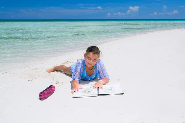 Charming pretty little girl, lying down on a sunny gorgeous beach and practicing  how to draw  in her sketch book — Φωτογραφία Αρχείου