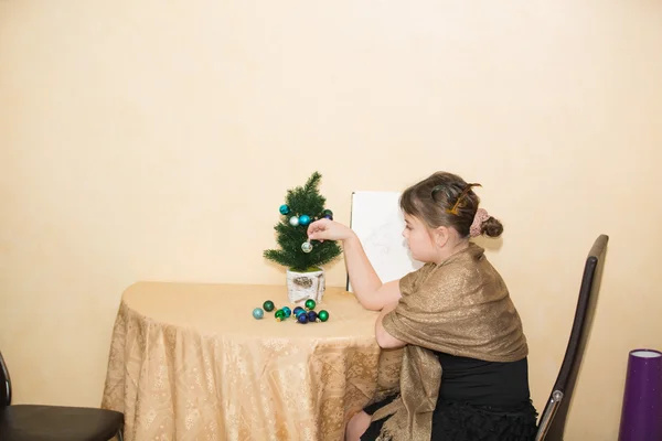 Маленькая девочка сидит одна за столом в своей комнате и украшает маленькую миниатюрную елку — стоковое фото