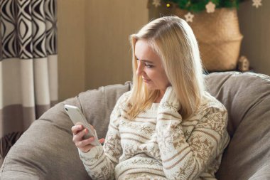 Bir kadın Noel kazağıyla bir sandalyede oturur ve Noel ağacının arkasındaki akıllı telefonuna bakar..