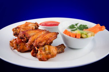 Kızarmış tavuk kanatları, havuç, kereviz ve sosla birlikte beyaz tabakta mavi arka planda..