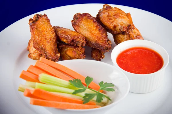 Roasted chicken wings with sticks carrots and celery and dipping sauce on white plate on blue background.