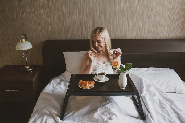 予期せぬ驚き ベッドで朝食 ベッドに座ってコーヒーを見ている女性の前にはテーブルトレイがあり クロワッサンと花瓶があります — ストック写真