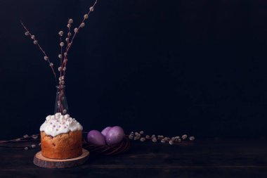 Karanlık bir arka planda paskalya pastaları, marşmelovlarla süslenmiş ve hasır bir yuvaya boyanmış Paskalya yumurtaları..