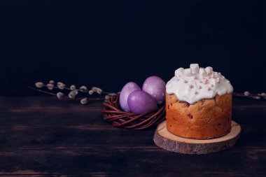 Karanlık bir arka planda paskalya pastaları, marşmelovlarla süslenmiş ve hasır bir yuvaya boyanmış Paskalya yumurtaları..