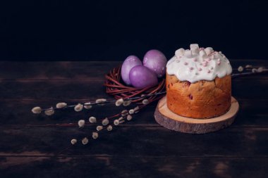 Karanlık bir arka planda paskalya pastaları, marşmelovlarla süslenmiş ve hasır bir yuvaya boyanmış Paskalya yumurtaları..