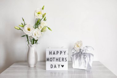 Furoshiki tekniğinde ipek kumaşa sarılı hediye kutusu, Anneler Günü 'nün kutlu olsun yazılı ışık kutusu, Eustoma' nın beyaz çiçekleri ya da ahşap arkaplanda Lisianthus vazosu. Sıfır Atık Yaşam Konsepti.