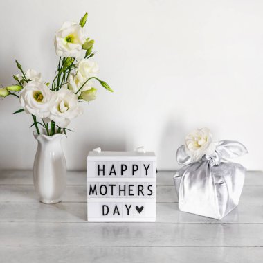 Furoshiki tekniğinde ipek kumaşa sarılı hediye kutusu, Anneler Günü 'nün kutlu olsun yazılı ışık kutusu, Eustoma' nın beyaz çiçekleri ya da ahşap arkaplanda Lisianthus vazosu. Sıfır Atık Yaşam Konsepti.