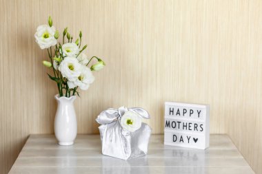 Furoshiki tekniğinde ipek kumaşa sarılı hediye kutusu, Anneler Günü 'nün kutlu olsun yazılı ışık kutusu, Eustoma' nın beyaz çiçekleri ya da ahşap arkaplanda Lisianthus vazosu. Sıfır Atık Yaşam Konsepti.