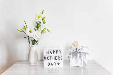 Furoshiki tekniğinde ipek kumaşa sarılı hediye kutusu, Anneler Günü 'nün kutlu olsun yazılı ışık kutusu, Eustoma' nın beyaz çiçekleri ya da ahşap arkaplanda Lisianthus vazosu. Sıfır Atık Yaşam Konsepti.