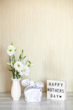 Furoshiki tekniğinde ipek kumaşa sarılı hediye kutusu, Anneler Günü 'nün kutlu olsun yazılı ışık kutusu, Eustoma' nın beyaz çiçekleri ya da ahşap arkaplanda Lisianthus vazosu. Sıfır Atık Yaşam Konsepti.