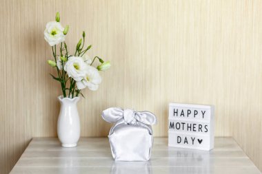 Furoshiki tekniğinde ipek kumaşa sarılı hediye kutusu, Anneler Günü 'nün kutlu olsun yazılı ışık kutusu, Eustoma' nın beyaz çiçekleri ya da ahşap arkaplanda Lisianthus vazosu. Sıfır Atık Yaşam Konsepti.