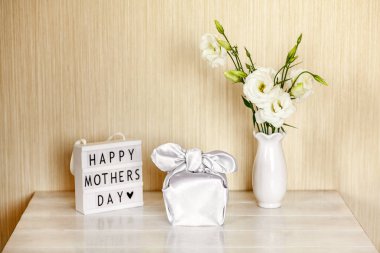 Furoshiki tekniğinde ipek kumaşa sarılı hediye kutusu, Anneler Günü 'nün kutlu olsun yazılı ışık kutusu, Eustoma' nın beyaz çiçekleri ya da ahşap arkaplanda Lisianthus vazosu. Sıfır Atık Yaşam Konsepti.