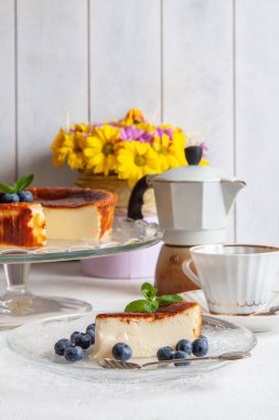 Ev yapımı Bask peynirli keki bir tabakta hafif arka planda yaban mersini ve nane yaprağı, bir fincan kahve, gayzer kahve makinesi, bir buket sarı çiçek..