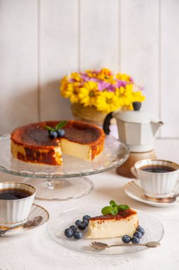 Ev yapımı ev yapımı Bask peynirli keki hafif arka planda yaban mersini ve nane yaprağı, bir fincan kahve, gayzer kahve makinesi, bir buket sarı çiçek..