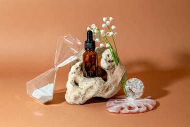 Sıra dışı bir doğal taşın arka planında serum şişesi olan modern, durgun yaşam, vadi zambakları ve kum saatleriyle dolu bir buket. Yaşlanma karşıtı bakım ürünleri kavramı.