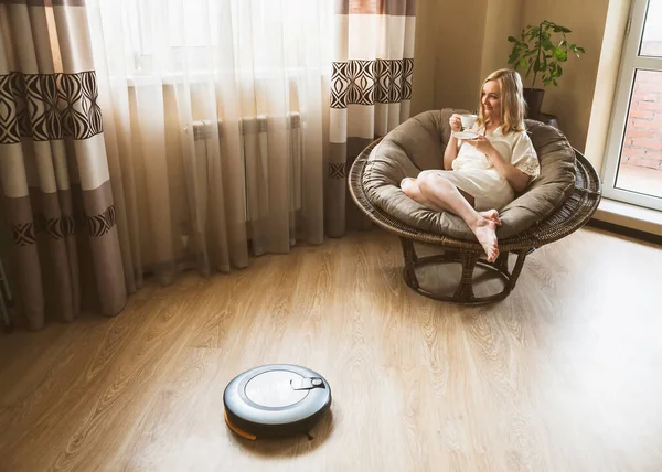 Robot elektrikli süpürge yeni dairesinde yerleri temizlerken mutlu kadın kahve içip dinleniyor. Ödev ve teknoloji kavramı.