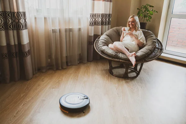 Robot elektrikli süpürge yeni dairesinde yerleri temizlerken mutlu kadın kahve içip dinleniyor. Ödev ve teknoloji kavramı.