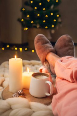 Kadın ayakları, ev kürkü terlikler, bir fincan kapuçino kalın iplik örtüsü üzerindeki mumlar ve ışıklı Noel ağacı. Umutsuzluk ve rahatlık atmosferi