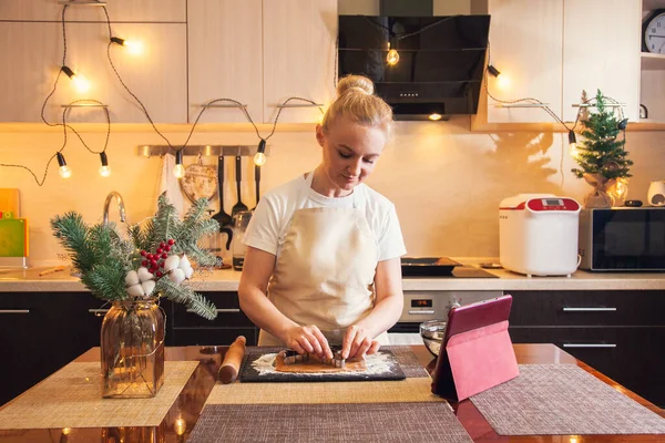 Woman following recipe on digital tablet and cuts out dough with mold for cooking christmas gingerbread cookies on her kitchen.