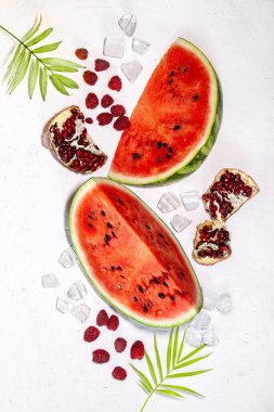 Yaz arkaplanı: Nar meyveli taze karpuz dilimleri, ahududu, buz, beyaz arka planda palmiye yaprağı, düz çizgi, üst manzara.