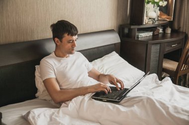 Orta yaşlı bir adam yatak odasında yatağında oturuyor ve bir dizüstü bilgisayar üzerinde çalışıyor. Uzaktan çalışma kavramı