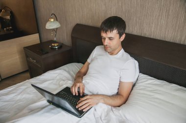 Orta yaşlı bir adam yatak odasında yatağında oturuyor ve bir dizüstü bilgisayar üzerinde çalışıyor. Uzaktan çalışma kavramı