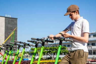 Genç bir adam akıllı telefonuyla QR kodunu tarayarak elektrikli scooter kiralıyor. Otoparkı paylaşırken mobiletleri tekmele..