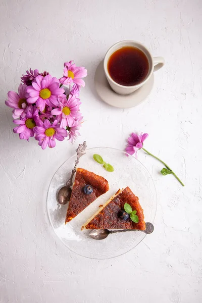 Slices of Homemade Basque burnt cheesecake on plate with blueberries and mint leaves, cup of black tea on light background with flowers in vase